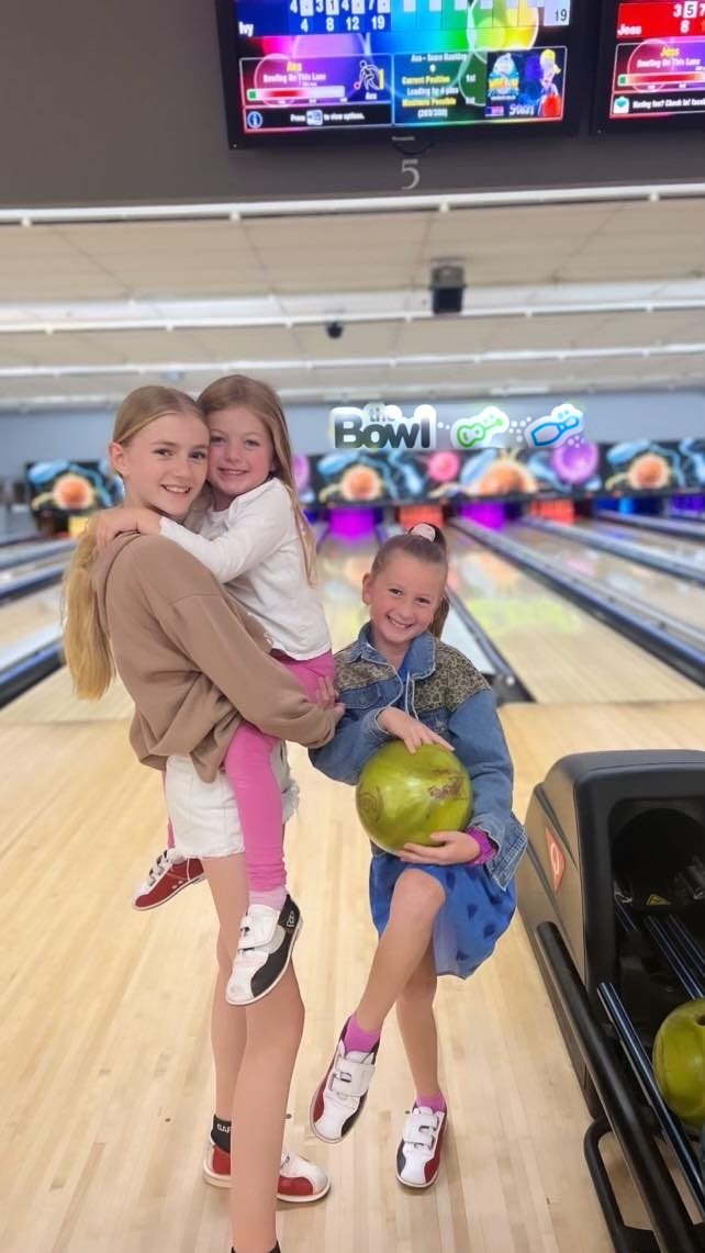 Three kids smiling in front of the bowling lanes at The Bowl, one holding a bowling ball
